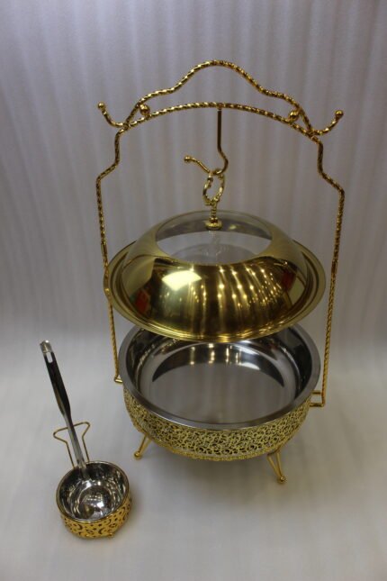 Luxury gold and stainless steel round chafing dish buffet server with a glass dome lid suspended on a gold frame, matching ladle, and buffet burner on a kitchen counter.