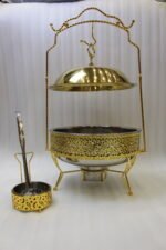 Luxury gold and stainless steel round chafing dish buffet server with a glass dome lid suspended on a gold frame, matching ladle, and buffet burner on a kitchen counter.Luxury gold and stainless steel round chafing dish buffet server with a glass dome lid suspended on a gold frame, matching ladle, and buffet burner on a kitchen counter.