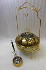 Luxury gold and stainless steel round chafing dish buffet server with a glass dome lid suspended on a gold frame, matching ladle, and buffet burner on a kitchen counter.