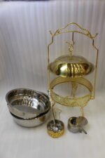 Luxury gold and stainless steel round chafing dish buffet server with a glass dome lid suspended on a gold frame, matching ladle, and buffet burner on a kitchen counter.