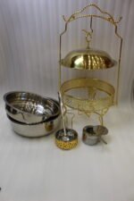 Luxury gold and stainless steel round chafing dish buffet server with a glass dome lid suspended on a gold frame, matching ladle, and buffet burner on a kitchen counter.