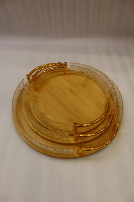 Set of three round wooden nesting serving trays with curved gold handles and clear textured glass rims stacked on a grey marble kitchen countertop.