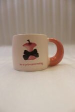 Cream ceramic coffee mug featuring a Snow White apple with a black bow, "Be a princess today" text, and a thick pink rounded handle sitting on a marble kitchen counter.Cream ceramic coffee mug featuring a Snow White apple with a black bow, "Be a princess today" text, and a thick pink rounded handle sitting on a marble kitchen counterCream ceramic coffee mug featuring a Snow White apple with a black bow, "Be a princess today" text, and a thick pink rounded handle sitting on a marble kitchen counter..
