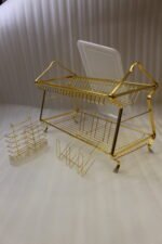 2 tier luxury gold metal dish drying rack with utensil basket, glass holder hooks, and clear drip tray on a kitchen countertop next to a faucet.