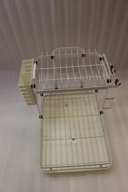 Cream two-tier metal dish drying rack with side utensil holder and drain tray on a kitchen countertop next to a sink.