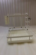 Cream two-tier metal dish drying rack with side utensil holder and drain tray on a kitchen countertop next to a sink.