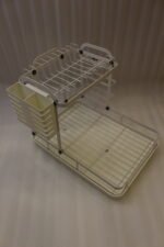 Cream two-tier metal dish drying rack with side utensil holder and drain tray on a kitchen countertop next to a sink.