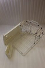Cream two-tier metal dish drying rack with side utensil holder and drain tray on a kitchen countertop next to a sink.