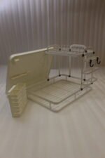 Cream two-tier metal dish drying rack with side utensil holder and drain tray on a kitchen countertop next to a sink.