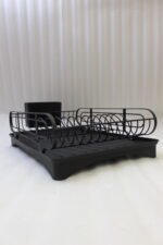 Matte black metal dish drying rack with attached utensil holder and drip tray sitting on a marble kitchen countertop beside a sink.