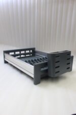 Modern grey and silver expandable kitchen dish drying rack with plate slots, utensil caddy, and drain spout sitting on a countertop next to a sink.
