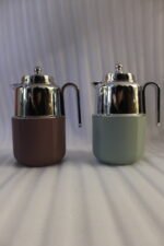 Two One More 1 Liter thermal vacuum coffee carafes in brown and sage green with ribbed bases and polished stainless steel tops on a kitchen counter.