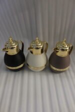Three luxury 1-liter thermal coffee and tea vacuum flasks with ornate gold lids and accents in matte black, cream, and taupe on a granite kitchen countertop.