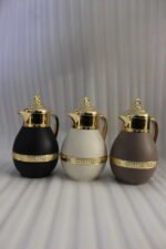 Three luxury 1-liter thermal coffee and tea vacuum flasks with ornate gold lids and accents in matte black, cream, and taupe on a granite kitchen countertop.